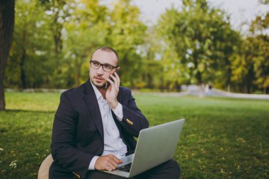 Genç işadamı beyaz gömlek, Klasik takım elbise, gözlük. Adam yumuşak kabarık üzerinde oturup, iş üstünde laptop pc bilgisayar, cep telefonu şehir Park açık havada yeşil çim üzerinde konuşmak. Mobil ofis, iş kavramı.