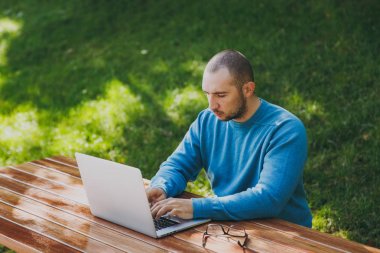 Genç başarılı akıllı adam işadamı veya öğrenci şehir Park açık havada yeşil doğa üzerinde çalışan dizüstü kullanarak cep telefonu ile masada oturan rahat mavi gömlekli gözlüklü. Mobil ofis kavramı.