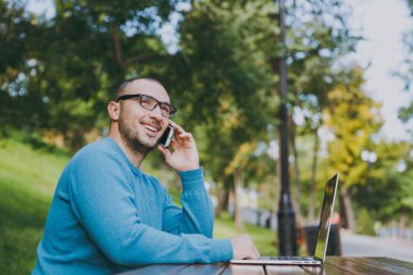 Genç akıllı adam işadamı veya öğrenci rahat mavi gömlekli masadan, gözlük gülümseyen başarılı cep telefonu, dizüstü bilgisayar kullanarak şehir Park açık havada çalışan konuşurken. Mobil ofis kavramı.