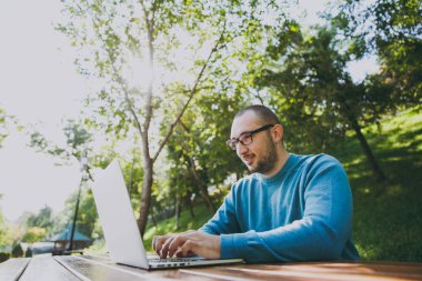 Genç başarılı akıllı adam işadamı veya öğrenci şehir Park açık havada yeşil doğa üzerinde çalışan dizüstü kullanarak cep telefonu ile masada oturan rahat mavi gömlekli gözlüklü. Mobil ofis kavramı.