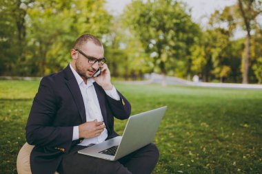 Genç işadamı beyaz gömlek, Klasik takım elbise, gözlük. Adam yumuşak kabarık üzerinde oturup, iş üstünde laptop pc bilgisayar, cep telefonu şehir Park açık havada yeşil çim üzerinde konuşmak. Mobil ofis, iş kavramı.