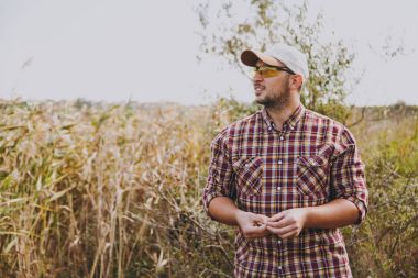 Tıraşsız delikanlı kareli gömlek, kap ve uzağa arıyorsunuz güneş gözlüğü solucan yemi güneşli bir günde çalılar ve sazlık arka planı balıkçılık için tutar. Yaşam tarzı, eğlence, eğlence kavramı