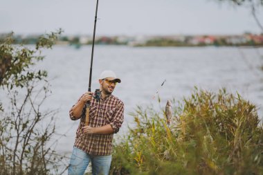 Tıraşsız delikanlı kareli gömlek, şapka ve güneş gözlüğü kutup yakalanan balık ile Balık tutma çekti ve çalılar ve sazlık yakınındaki göl kıyısında sevinir. Yaşam tarzı, rekreasyon, balıkçı eğlence kavramı