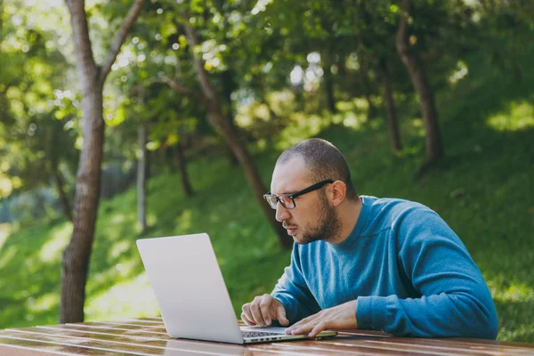 Young successful smart man businessman or student in casual blue shirt glasses sitting at table with mobile phone in city park using laptop working outdoors on green nature. Mobile Office concept.