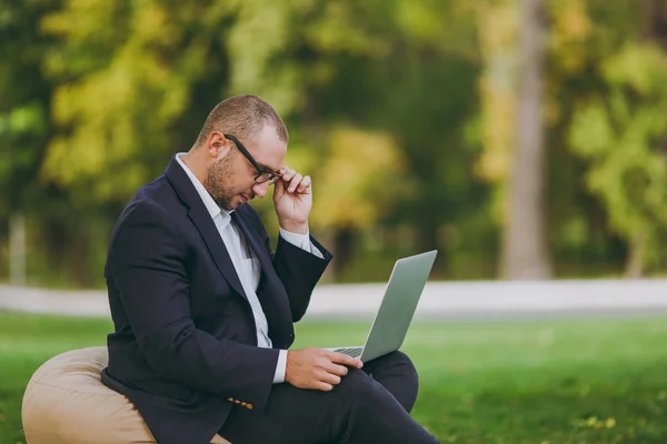 Молодой успешный бизнесмен в белой рубашке, классическом костюме, очках. Человек сидит на мягком пуфе, работая на ноутбуке компьютера в городском парке на зеленой лужайке на природе. Мобильный офис, концепция бизнеса . — стоковое фото