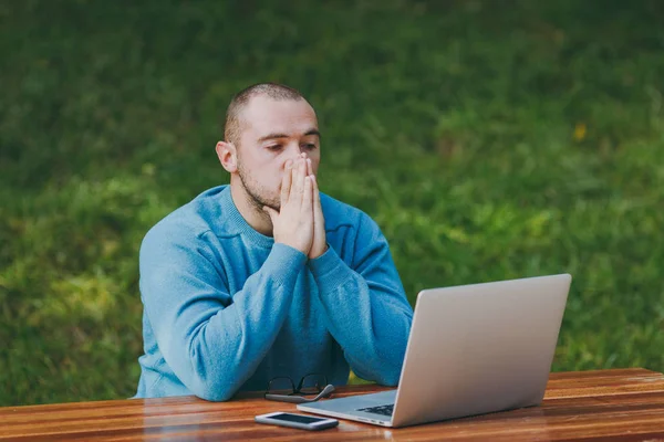 Upset sad disturb man businessman or student in casual blue shirt, sitting at table in city park using laptop working outdoors, hold hands at his face, concerned about problems. Mobile Office concept.