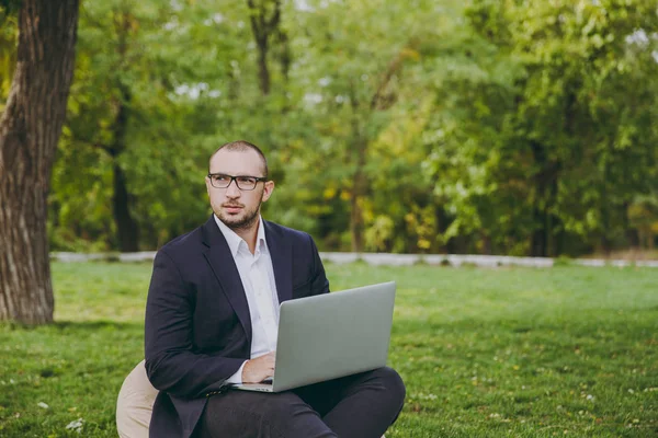 Młodym biznesmenem w klasyczny garnitur, biała koszula, okulary. Człowiek siedzieć na miękką pufę, pracując na komputerze pc laptop w parku miejskim na zielony trawnik na zewnątrz na charakter. Office Mobile, koncepcja biznesowa. — Zdjęcie stockowe