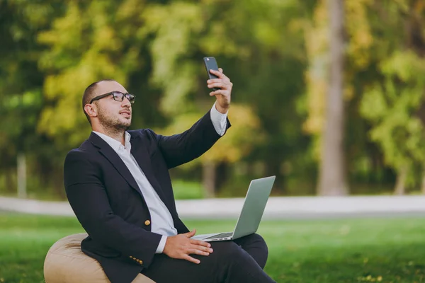 Młody biznesmen w klasyczny garnitur, biała koszula, okulary. Człowiek siedzieć na miękką pufę z komputerem pc laptop, robić selfie na telefon komórkowy w parku miejskim na zielony trawnik na zewnątrz. Koncepcja biznesowa Mobile Office. — Zdjęcie stockowe