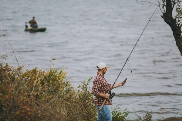 Jovem homem sem barba em camisa quadriculada, boné e óculos de sol puxado para fora pólo de pesca e detém peixes capturados na costa do lago perto juncos contra fundo de barco. Estilo de vida, conceito de lazer do pescador — Fotografia de Stock