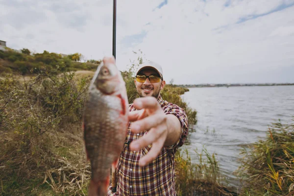 Jonge ongeschoren man in geruite shirt, pet, zonnebril houdt van hengel en strekt zich uit zijn hand tot gevangen vis op de oever van het meer in de buurt van struiken en riet. Lifestyle, recreatie, visser leisure concept — Stockfoto