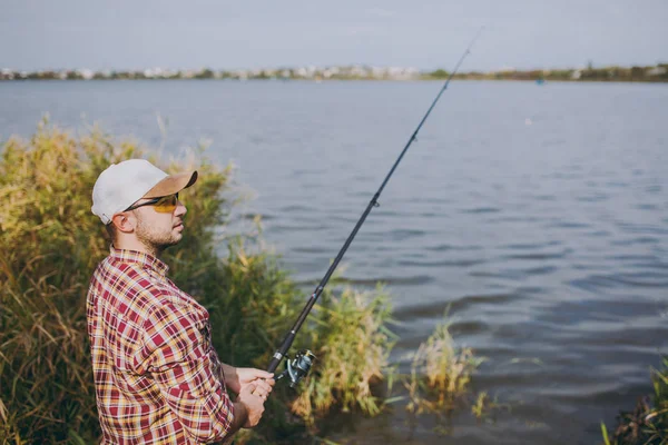 Visão lateral Jovem homem sem barba com uma vara de pesca em camisa quadriculada, boné e óculos de sol olha para a distância no lago da costa perto de arbustos e juncos. Estilo de vida, recreação, conceito de lazer do pescador . — Fotografia de Stock