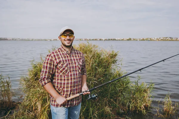 Joven hombre sonriente sin afeitar en camisa a cuadros, gorra y gafas de sol sostiene caña de pescar en la orilla del lago en el fondo de agua, arbustos y cañas. Estilo de vida, recreación, concepto de ocio del pescador . —  Fotos de Stock