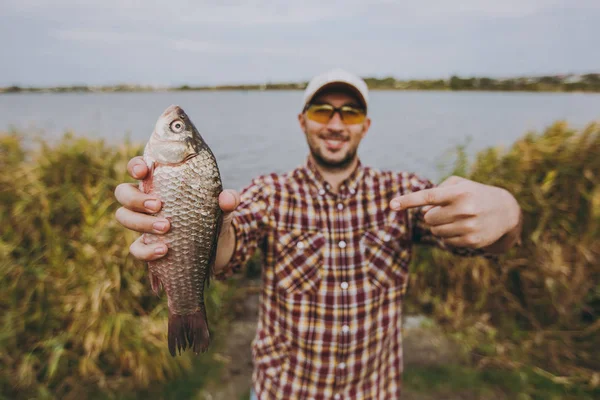 Joven hombre sin afeitar en camisa a cuadros, gorra y gafas de sol pescado capturado, lo muestra y señala con un dedo en ella en la orilla del lago en el fondo del agua y cañas. Estilo de vida, concepto de ocio del pescador —  Fotos de Stock