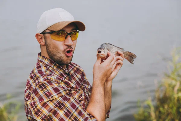 Yakın genç kareli gömlek, şapka ve güneş gözlüğü tıraşsız erkekte bir balık yakaladık, o göstermek ve su arka plan üzerinde göl kıyısında sevinir. Yaşam tarzı, rekreasyon, balıkçı eğlence kavramı — Stok fotoğraf