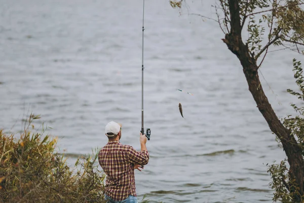 Vista posteriore Giovane uomo non rasato con una canna da pesca in camicia a scacchi e cappuccio tira fuori canna da pesca con i pesci catturati sul lago dalla riva vicino ad arbusti e canne. Stile di vita, concetto di tempo libero del pescatore . — Foto Stock