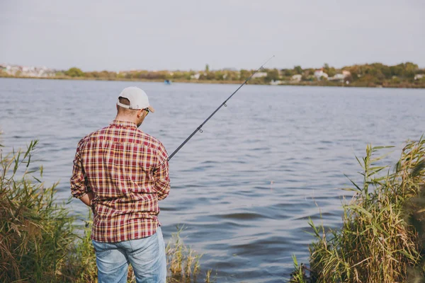 ΠΙΣΩ ΟΨΗ αξύριστος νεαρός με καλάμι ψαρέματος στο καρώ πουκάμισο, καπάκι και γυαλιά ηλίου ρίχνει δόλωμα και το ψάρεμα στη λίμνη από την ακτή κοντά σε θάμνους και τα καλάμια. Lifestyle, ψυχαγωγία, αναψυχής έννοια ψαράς. — Φωτογραφία Αρχείου