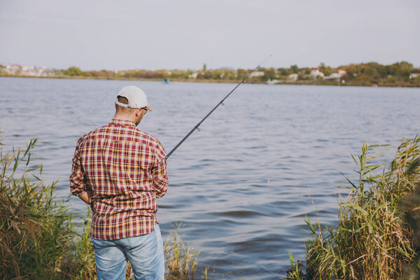 Back view Young unshaven man with fishing rod in checkered shirt, cap and sunglasses casts bait and fishing on lake from shore near shrubs and reeds. Lifestyle, recreation, fisherman leisure concept.