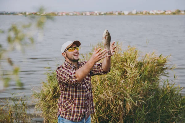 Młody ogolony uśmiechnięty człowiek w kraciaste koszule, cap, okulary złowionych ryb i rzucić się na brzegu jeziora na tle wody, krzewów i trzciny. Styl życia, rekreacja, koncepcja wypoczynek rybak — Zdjęcie stockowe