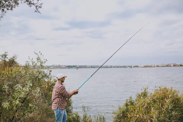 Visão lateral Jovem homem sem barba com uma vara de pesca em camisa quadriculada, boné e óculos de sol lança um pólo de pesca no lago de costa perto de arbustos e juncos. Estilo de vida, recreação, conceito de lazer do pescador — Fotografia de Stock