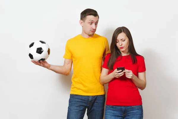 Fun engaged young man, indifferent woman send sms on mobile phone, football fans cheer up support team with soccer ball isolated on white background. Sport, couple, family leisure, lifestyle concept.