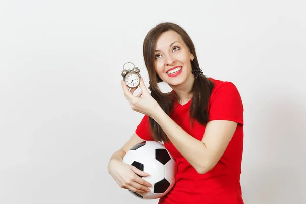 European young woman, two fun pony tails, football fan or player in red uniform hold old alarm clock, soccer ball isolated on white background. Sport play football health, time lifestyle concept.