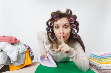 Eğlenceli yakın curlers Ütü Masası ile demir aile giyim standında hafif giysiler içinde saç ile ev hanımı. Dudaklar shhh jest beyaz arka plan üzerinde izole parmağında ile kadın söyleyerek sus sus.