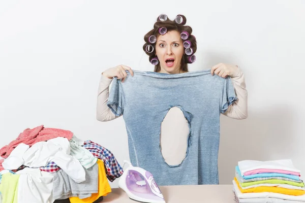 Scioccato divertimento folle casalinga, bigodini sui capelli in abiti leggeri in possesso di camicia bruciata con foro fatto da ferro, in piedi a asse da stiro. Donna isolata su sfondo bianco. Copia spazio per pubblicità . — Foto Stock