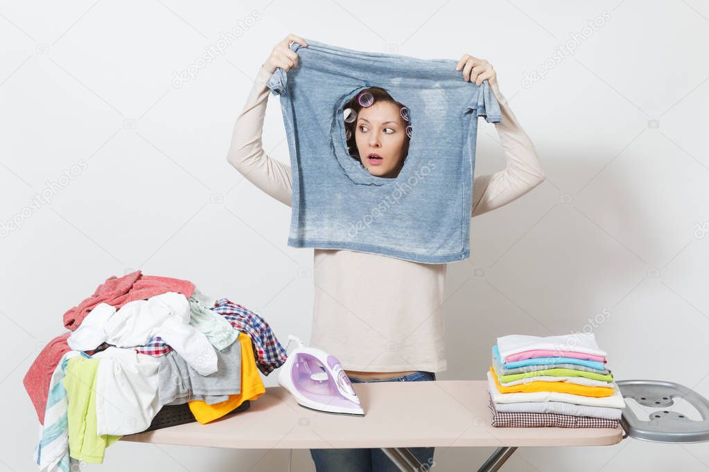 Shocked housewife, curlers on hair in light clothes holding burned shirt, looking through hole made by iron standing at ironing board. Woman isolated on white background. Copy space for advertisement.