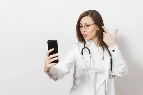 Sonriente divertida hermosa mujer doctora joven con estetoscopio, gafas aisladas sobre fondo blanco. Doctora en bata médica haciendo selfie en el teléfono móvil. Personal sanitario, salud, medicina — Foto de Stock