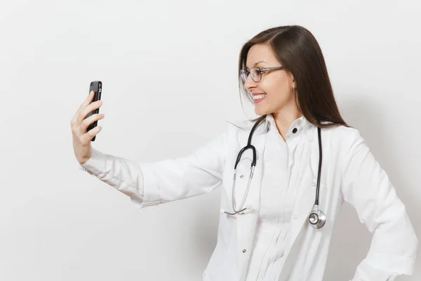 Sonriente divertida hermosa mujer doctora joven con estetoscopio, gafas aisladas sobre fondo blanco. Doctora en bata médica haciendo selfie en el teléfono móvil. Personal sanitario, salud, medicina — Foto de Stock