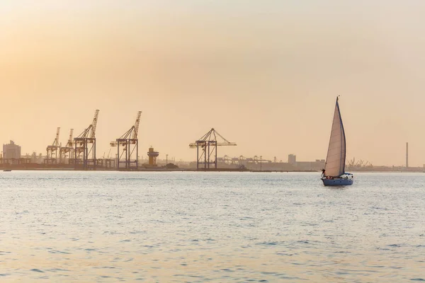 Beautiful seascape. Lonely boat with sails in a calm sea with reflection of sunlight near a industrial seaport on the coastline against the background of a clear warm evening sunset