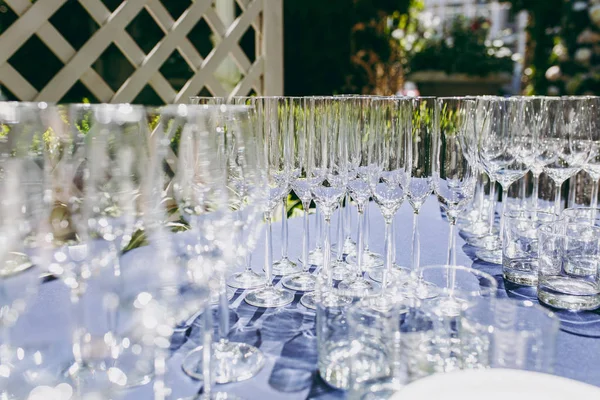 Preparing for party. Many clean shiny empty glasses for wine, champagne, whiskey, juice, water and other drinks on the table with a blue tablecloth in the gazebo at an outdoor party in the garden — Stock Photo, Image