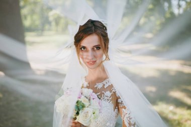 Güzel düğün photosession. Beyaz dantel elbise ve weathery güneşli günde büyük bir yeşil bahçe içinde çırpınan bir peçe beyaz ve pembe çiçekler buket genç gülümseyen gelin portresi