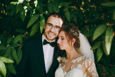 Güzel düğün photosession. Siyah takım elbise, beyaz gömlek, papyon ve gözlük ve gelin peçe ile zarif dantel elbise ve dekorasyon dallar yeşil Bush ortasında saçında damat