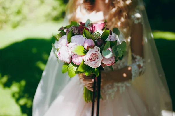 Närbild av en vacker anbud brudbukett i rosa färger av rosor, pioner och gröna kvistar bundna med en lång sammet rödvin menyfliksområdet i händerna på bruden. Bröllop tillbehör för bruden — Stockfoto