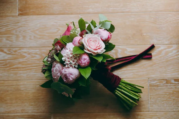Nahaufnahme eines wunderschönen zarten Brautstraußes in rosafarbenen Farben von Rosen, Pfingstrosen und grünen Zweigen, der mit einem langen samtenen weinroten Band auf einem Holzboden gebunden ist. Hochzeitsaccessoires für Braut — Stockfoto