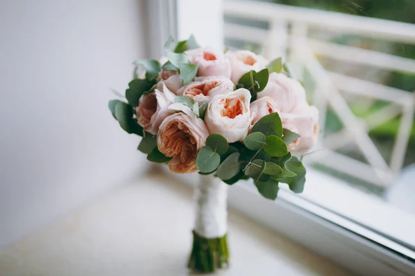 Close Um lindo buquê de noiva macio em rosas em forma de pião e galhos verdes amarrados com uma longa fita branca de seda em um peitoril bege. Acessórios de casamento para noiva — Fotografia de Stock