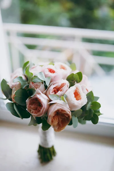 Schließen Sie einen wunderschönen zarten Brautstrauß in rosafarbenen Farben aus pionenförmigen Rosen und grünen Zweigen, die mit einem langen seidenweißen Band auf einer beigen Fensterbank gebunden sind. Hochzeitsaccessoires für Braut — Stockfoto