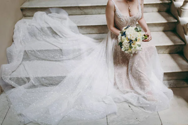 Hermosa celebración de boda. La novia en elegante vestido de novia de encaje de marfil con un collar y un ramo de peonías y bayas azules se sienta en escalones de piedra que se extienden falda. Ropa de boda, accesorios — Foto de Stock