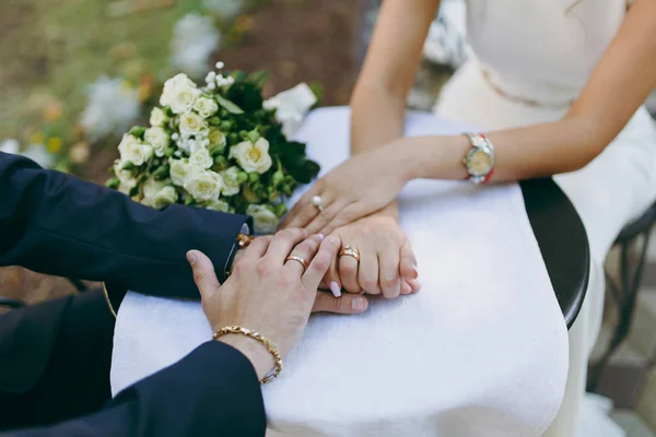 Bela celebração de casamento. O noivo de terno com pulseira e anel de casamento e a noiva em vestido de marfim com relógio na mão e buquê de rosas brancas de mãos dadas na mesa com toalha de mesa — Fotografia de Stock