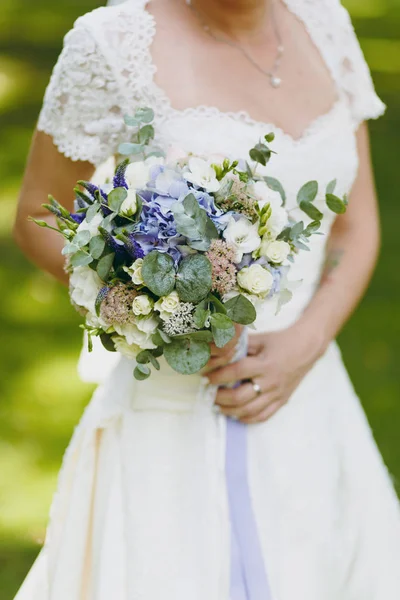 Bela celebração de casamento. Um buquê de várias flores leves, folhas verdes e galhos com uma fita roxa nas mãos da noiva em um elegante vestido de renda com mangas curtas no jardim — Fotografia de Stock