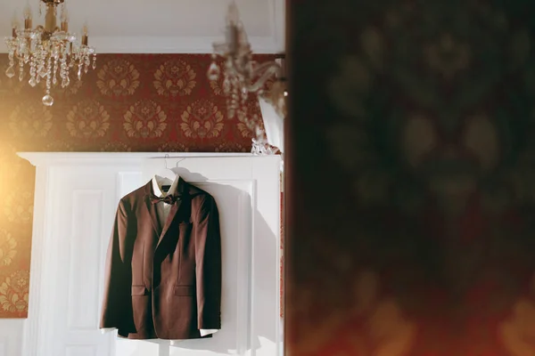 Hermosa chaqueta a cuadros marrón boda, una camisa blanca y una corbata para el novio cuelgan de los hombros en la puerta de la habitación con hermoso fondo de pantalla con dibujos. Ropa de boda, accesorios — Foto de Stock