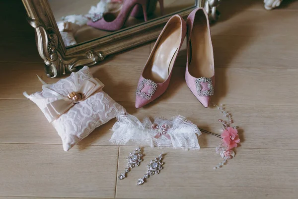 Close up Elegant light pink wedding textile shoes with decorations, pillow with rings, earrings, garter and hairpin for the bride on wooden floor background near the mirror. Wedding wear, accessories — Stock Photo, Image