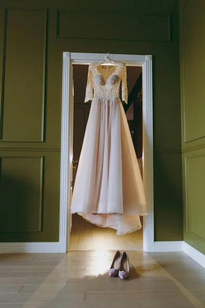 Um lindo e elegante vestido de noiva com rendas e mangas para a noiva pendura em um cabide na entrada sobre sapatos de casamento rosa na sala verde. Casamento desgaste, acessórios — Fotografia de Stock