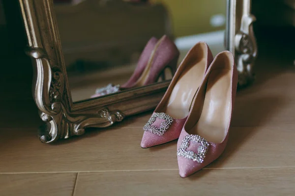 Cerrar Elegantes zapatos brillantes textiles de boda rosa claro con grandes decoraciones en las narices afiladas para la novia en el fondo del piso de madera cerca del espejo en un hermoso marco. Ropa de boda, accesorios — Foto de Stock