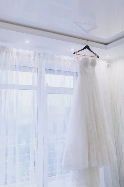 Um lindo e elegante vestido de noiva com rendas e mangas curtas para a noiva está pendurado em um cabide contra o fundo de uma grande janela com tule branco. Casamento desgaste, acessórios — Fotografia de Stock