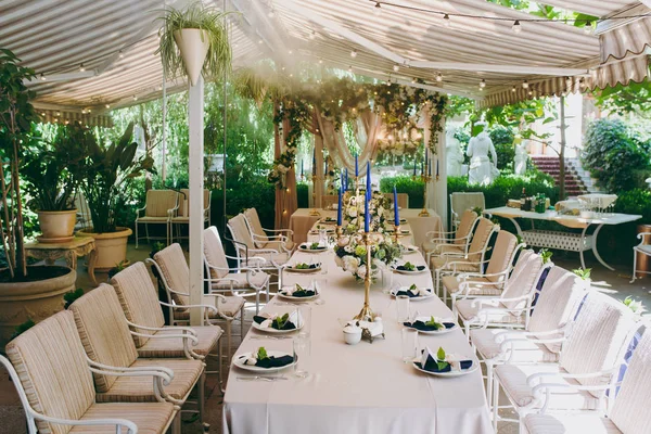 Bonita e requintada decoração da celebração do casamento no meio de um jardim verde. Banquete mesa servida com uma toalha de mesa bege, pratos e castiçais com velas azuis sob cobertura — Fotografia de Stock