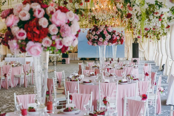 Mooie decoratie van de feestzaal huwelijk onder de luifel in roze, donkerrood en witte tinten. Hall met tafels en stoelen versierd met dunne doek, boeketten, bloemenslingers en confetti — Stockfoto