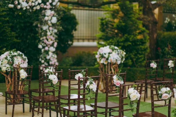 Schöne Dekoration einer Hochzeitszeremonie in einem grünen herbstlichen Garten. Braune Holzstühle für Gäste auf beiden Seiten des Gehwegs, der zu einem festlichen Bogen führt, der mit Blumen geschmückt ist — Stockfoto