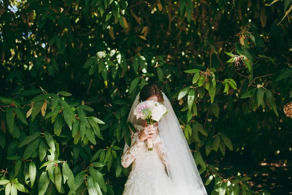 Beautiful wedding photosession. Young tender cute bride in elegant dress with long sleeves and veil covers her face with bouquet of white and pink flowers amidst the branches of green bush in garden — Stock Photo, Image